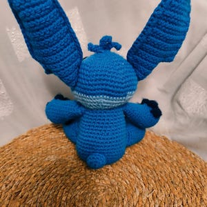 May include: A handmade blue crochet doll with large ears, small arms, and a small tail. The doll is sitting on a woven, circular mat. The doll's body is a solid blue, with lighter blue accents around the neck and on the ears.