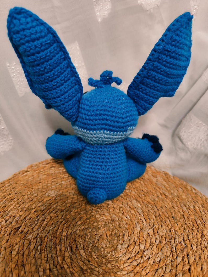 May include: A handmade blue crochet doll with large ears, small arms, and a small tail. The doll is sitting on a woven, circular mat. The doll's body is a solid blue, with lighter blue accents around the neck and on the ears.
