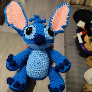 May include: A handmade blue crochet Stitch doll with large ears, black eyes, and a small nose. The ears have a pink interior. The doll has dark blue accents on its feet and hands. The doll is approximately 15 cm tall.