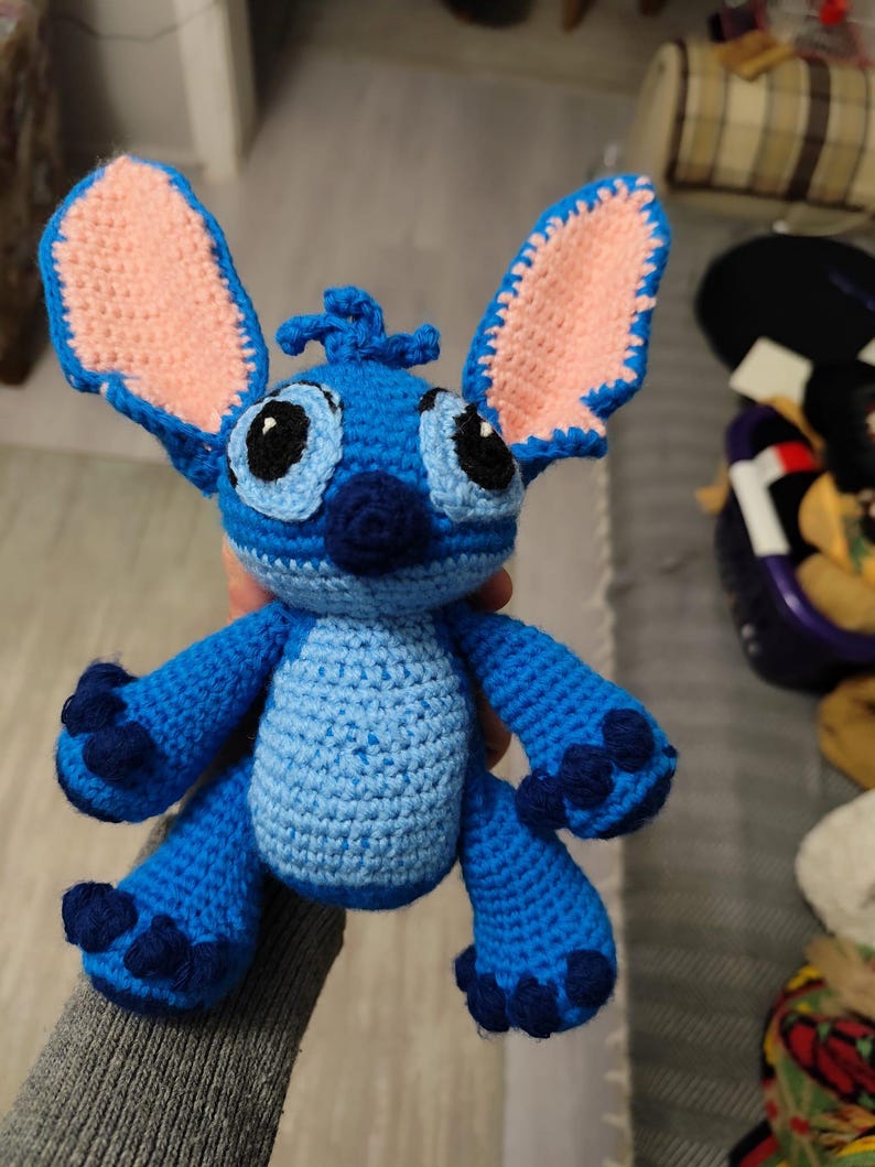 May include: A handmade blue crochet Stitch doll with large ears, black eyes, and a small nose. The ears have a pink interior. The doll has dark blue accents on its feet and hands. The doll is approximately 15 cm tall.