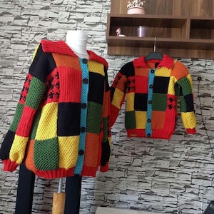 Puede incluir: Un cárdigan patchwork colorido y un cárdigan a juego para niños. Los cárdigans presentan una mezcla de cuadrados rojos, amarillos, verdes, negros y naranjas. El cárdigan para adultos se exhibe en un maniquí. El cárdigan para niños está en una percha.