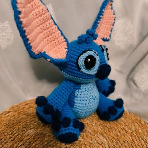 May include: A handmade blue crochet Stitch doll with large ears, a light blue belly, and dark blue accents. The ears have pink inner details. The doll is sitting on a woven, circular surface.