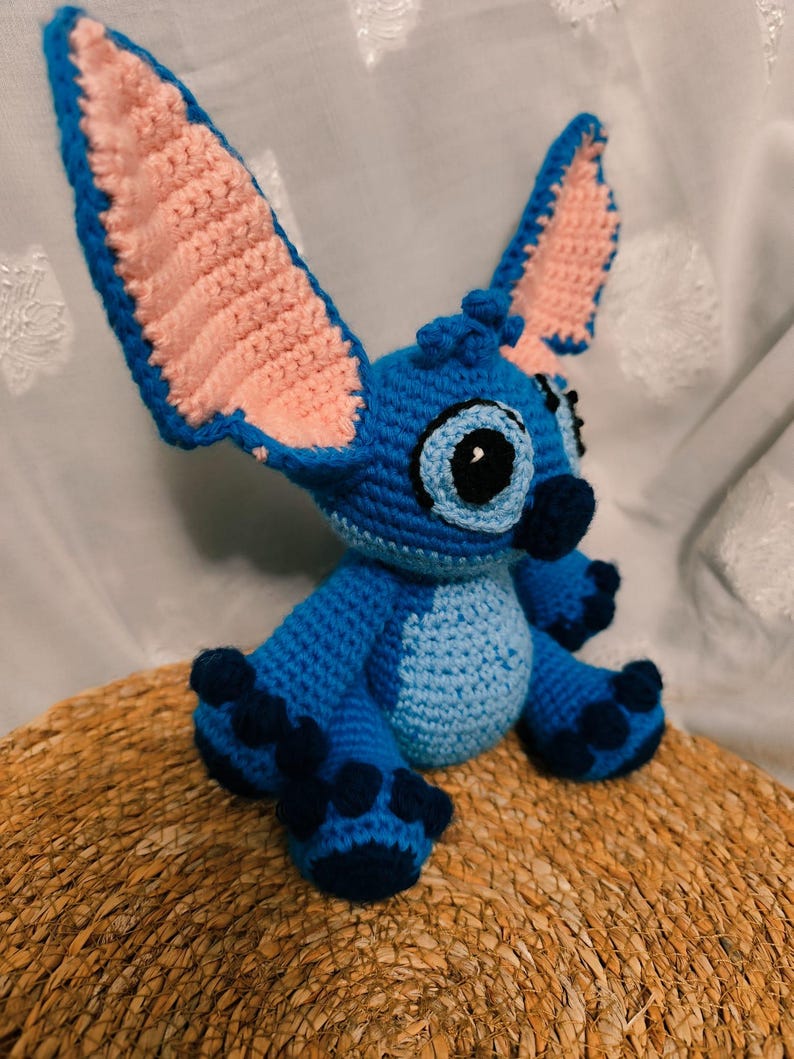 May include: A handmade blue crochet Stitch doll with large ears, a light blue belly, and dark blue accents. The ears have pink inner details. The doll is sitting on a woven, circular surface.