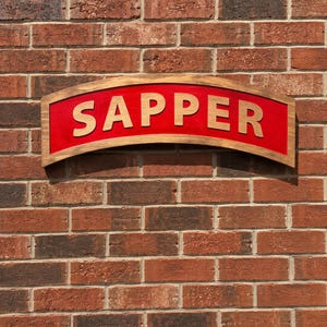 May include: A curved wooden sign with a dark brown stained frame and a bright red center panel. The word SAPPER is displayed in large, raised, light-colored wooden capital letters across the center of the red background of the sign.