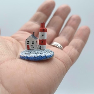 May include: A miniature lighthouse and house painted on a small wooden base. The lighthouse is red and white with a black stripe. The house is white with a red door and black windows. The base is painted blue and white to resemble water.