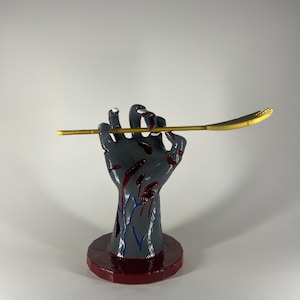 May include: A grey hand sculpture with red blood-like splatters and blue vein details, mounted on a circular dark red base. The hand is holding a long, thin, metallic gold-coloured rod that extends horizontally across the fingers.