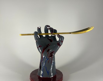 Pode incluir: Uma escultura de mão cinzenta com salpicos vermelhos semelhantes a sangue e detalhes de veias azuis, montada numa base circular vermelho-escura. A mão segura uma vareta longa e fina de cor dourada metálica que se estende horizontalmente.