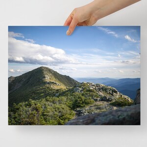 May include: A panoramic view from a mountaintop, showing a lush green landscape with rocky outcroppings. The sky is a bright blue with fluffy white clouds.