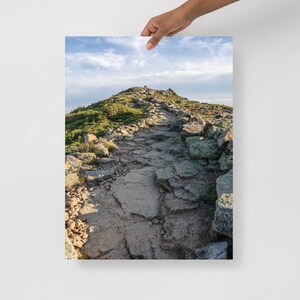 May include: A rocky path winds its way up a mountainside, leading to a peak in the distance. The path is lined with green vegetation and large rocks, and the sky is a bright blue with white clouds.