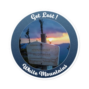May include: A blue and white round sticker with the text "Get Lost!" and "White Mountains" in a white cursive font. The sticker features a photo of a wooden sign that reads "Entering Pemigewasset Wilderness White Mountain National Forest" with a sunset in the background.