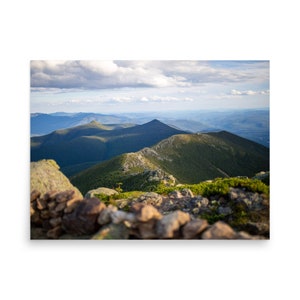 May include: A panoramic view from a mountaintop, showcasing rolling green hills and a distant valley. The foreground features a rocky outcropping covered in green vegetation.