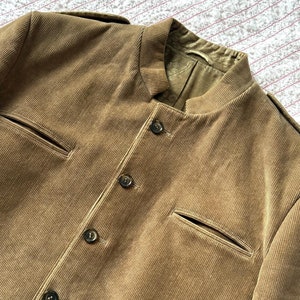 Puede incluir: Una chaqueta de pana marrón con cuello mandarín y cierre de botones. La chaqueta tiene dos bolsillos en la parte delantera y un interior forrado.