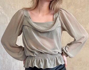 May include: A light olive green blouse with long, sheer sleeves and a square neckline. The blouse has a gathered waist and a ruffled hem. The model is wearing black leather pants.