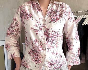 May include: A button-down shirt with a floral pattern in shades of pink and purple on a cream background. The shirt is paired with vertically striped, wide-leg pants in shades of gray and white. The outfit is casual and stylish.