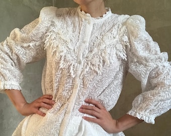 May include: A white cotton blouse with a lace trim and embroidered details. The blouse has a high neck and long sleeves with ruffles at the cuffs. The blouse is cinched at the waist and has a flowing skirt.