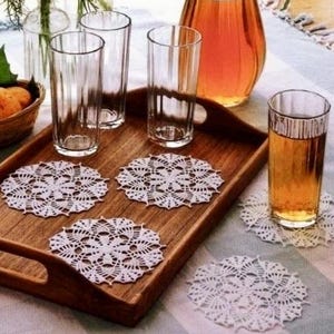 Puede incluir: Una bandeja de madera con cuatro vasos transparentes, una jarra de líquido ámbar y un vaso de líquido ámbar. Los posavasos de encaje blanco están debajo de los vasos. También hay una cesta de pan y un jarrón con flores.