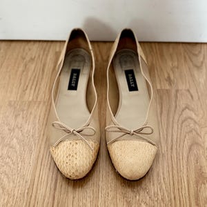 Op de afbeelding: Een paar beige ballerina's met een slangenhuidpatroon op de neus. De ballerina's hebben een strik aan de voorkant en het woord "BALLY" staat aan de binnenkant van elke schoen.