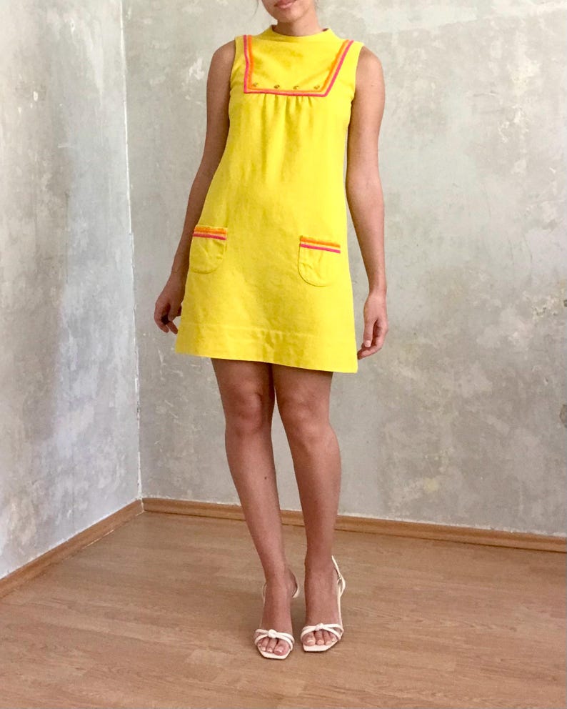 May include: A sleeveless yellow mini dress with a high neckline and orange and pink trim. The dress has two pockets on the front and is made of a soft, textured fabric.