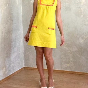 May include: A sleeveless yellow mini dress with a high neckline and a contrasting orange and pink trim. The dress has two pockets on the front and a row of buttons down the center.