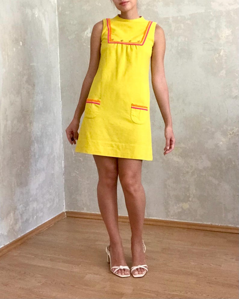 May include: A sleeveless yellow mini dress with a high neckline and a contrasting orange and pink trim. The dress has two pockets on the front and a row of buttons down the center.