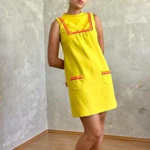 May include: A sleeveless yellow mini dress with a high neckline and colorful trim. The dress has two pockets with colorful trim on the front.