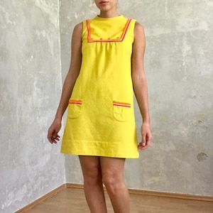 May include: A sleeveless yellow mini dress with a high neckline and contrasting pink and orange trim. The dress has two pockets on the front and a slightly flared skirt.
