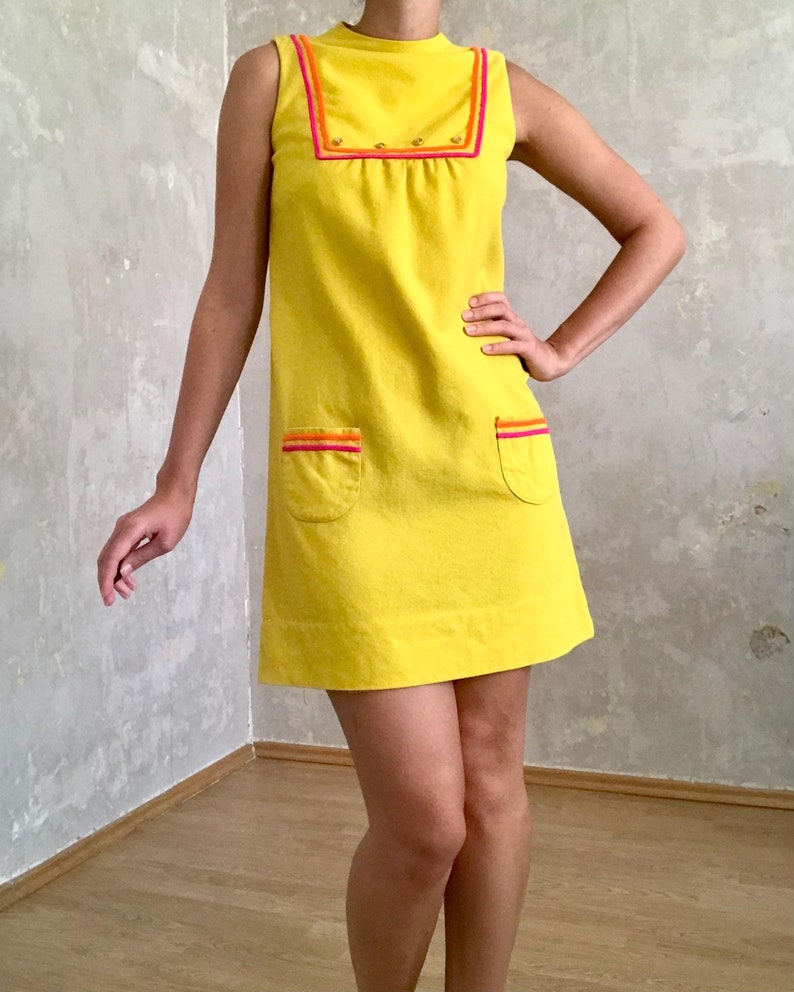 May include: A sleeveless yellow mini dress with a high neckline and a contrasting pink and orange trim. The dress has two pockets on the front and a row of gold buttons on the neckline.