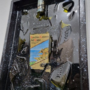 Puede incluir: Una obra de arte enmarcada en negro con una botella de vidrio verde rota en pedazos y pegada a la superficie negra. La etiqueta de la botella dice "Vecchio Amaro del Capo" y "Liquore d'Erbe di Calabria". El fondo es negro con motas blancas.