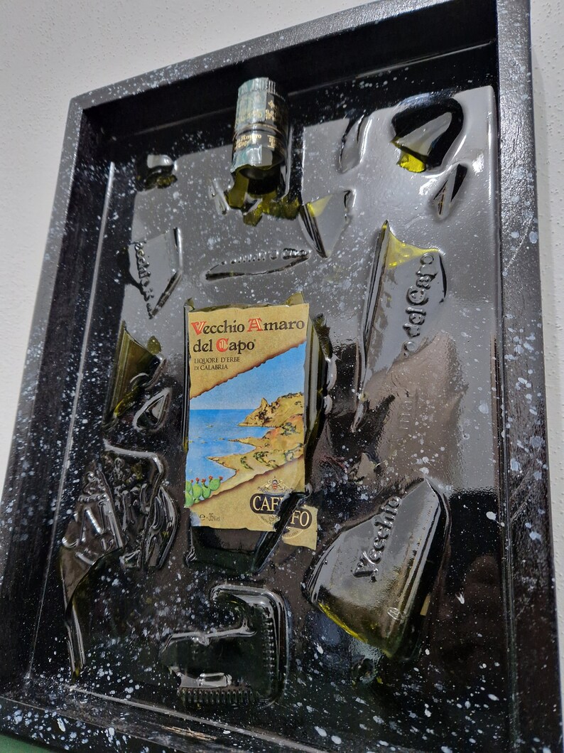 Puede incluir: Una obra de arte enmarcada en negro con una botella de vidrio verde rota en pedazos y pegada a la superficie negra. La etiqueta de la botella dice "Vecchio Amaro del Capo" y "Liquore d'Erbe di Calabria". El fondo es negro con motas blancas.