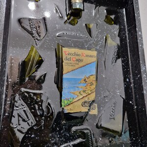 Puede incluir: Un marco negro con una resina transparente llena de trozos rotos de una botella de vidrio verde. La etiqueta de la botella dice "Vecchio Amaro del Capo" y "Liquore d'Erbe di Calabria".