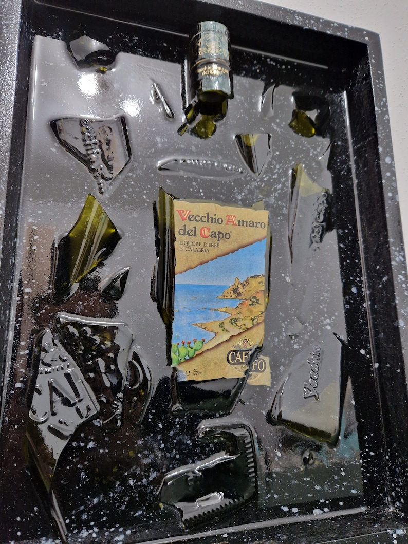Puede incluir: Un marco negro con una resina transparente llena de trozos rotos de una botella de vidrio verde. La etiqueta de la botella dice "Vecchio Amaro del Capo" y "Liquore d'Erbe di Calabria".