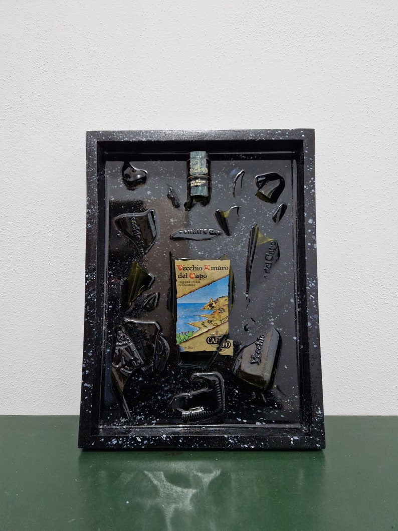 Puede incluir: Una imagen enmarcada en negro con un fondo moteado de blanco. Dentro del marco hay varios trozos de vidrio roto, incluida una botella de licor "Vecchio Amaro del Capo".