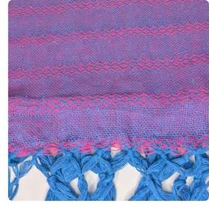 May include: A woven textile with a vibrant color palette. The fabric features a purple base with horizontal pink and blue patterned stripes. The bottom edge is adorned with a fringe of bright blue knotted tassels, adding a decorative element.
