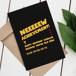 May include: A black birthday card with yellow text that says "NEEEEEW ACHIEVEMENT!" and "HAPPY BIRTHAY CRAWLER EWWWW YOU'RE OLD NOW." The card is on a brown envelope with pink splatter accents.