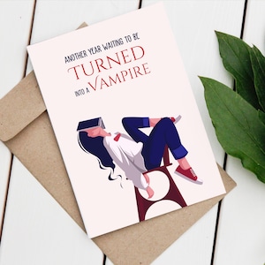 May include: A white card with red and blue text that reads "Another year waiting to be turned into a vampire". A cartoon illustration of a person sitting on a chair with a book over their head.