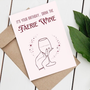 May include: A pink birthday card with the text "It's your birthday... Drink the Faerie Wine" in maroon lettering. A hand holding a wine glass with a maroon outline is surrounded by a circle of maroon stars.