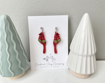 May include: A pair of red cardinal earrings with green holly leaves and red berries. The earrings are handmade and are on a white card with the text "Cardinal Clay Company Handmade Clay Jewelry".