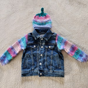 May include: A blue denim jacket with rainbow striped knit sleeves and a pink, blue, and green knit hat with a green pom-pom.