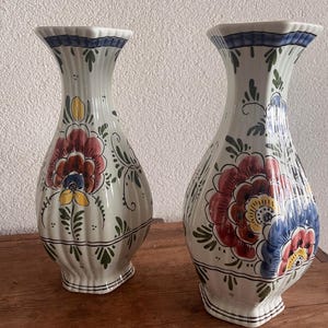 May include: Two white ceramic vases with a vertical ribbed texture and a flared rim. Each vase features hand-painted floral motifs in red, blue, yellow, and green, with a blue band around the top rim and a small base at the bottom.