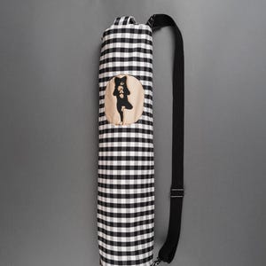 May include: A yoga mat bag in a black and white checkered pattern. The bag has a black shoulder strap and a circular patch with a black silhouette of a person in a yoga pose. The bag is designed to carry a yoga mat.