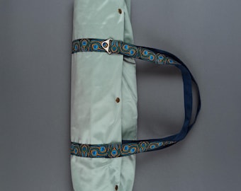 May include: A rolled-up, light green yoga mat secured with a patterned strap and a navy blue handle. The strap features a repeating design of blue and gold circular patterns. The mat is fastened with brown buttons.