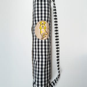 May include: A yoga mat bag with a black and white gingham pattern. The bag features a tan patch with a yellow cartoon dog in a yoga pose. The bag has a matching black and white gingham strap.