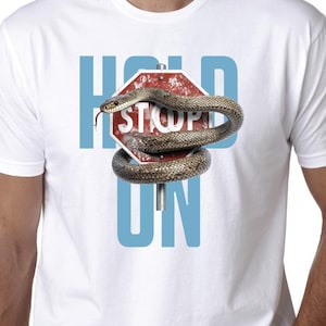 May include: White t-shirt featuring a graphic design. The design includes a red stop sign with a snake wrapped around it, and the words "HOLD ON" in blue block letters. The snake is brown and tan.