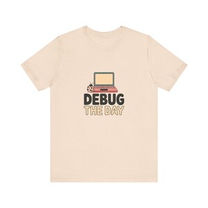 May include: A light beige short-sleeve t-shirt featuring a graphic of a red laptop with a small ladybug next to it. Below the laptop, the text reads DEBUG THE DAY in bold, dark letters. The design is centered on the chest of the garment.