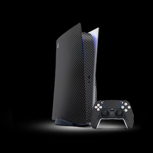 Puede incluir: Una consola PlayStation 5 negra y blanca con un diseño de fibra de carbono. La consola está abierta y la unidad de disco es visible. También se incluye un mando PlayStation 5 negro y blanco.