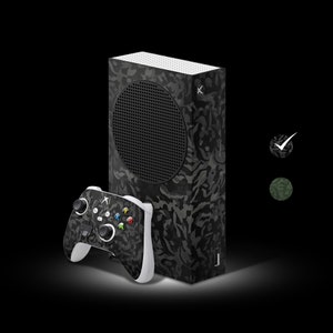 May include: A black and gray camouflage Xbox Series X console with a matching controller. The console has a black disc drive and a USB port on the front. The controller has a black and gray camouflage design with white buttons.