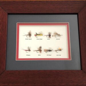 May include: A framed display of eight different fly fishing lures, each with a unique design and color scheme. The lures are mounted on a white background with their names printed below them. The frame is made of dark wood with a red and black mat.