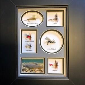 May include: A framed display of six different fly fishing lures, each with a unique design and name. The lures are displayed on a gray background with a black frame. The names of the lures are "Robert's Yellow Drake", "Adams", "Michigan Hillbilly", "Au Sable Shunk", "North Branch", and "Professor".