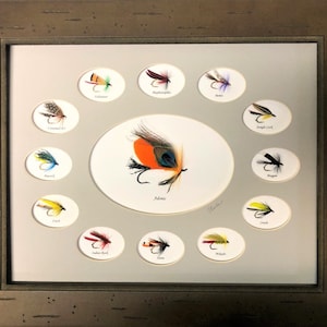 May include: A framed display of 12 different fly fishing lures, each mounted in a separate oval-shaped frame. The lures are arranged in a circle around a larger oval frame containing a single lure labeled "Adams".