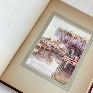 May include: An open book with a framed watercolor print of a Japanese scene. The print depicts a wooden bridge over water, with people and wisteria flowers. The book's pages are aged, and the print is titled "Wistaria at Kameda".